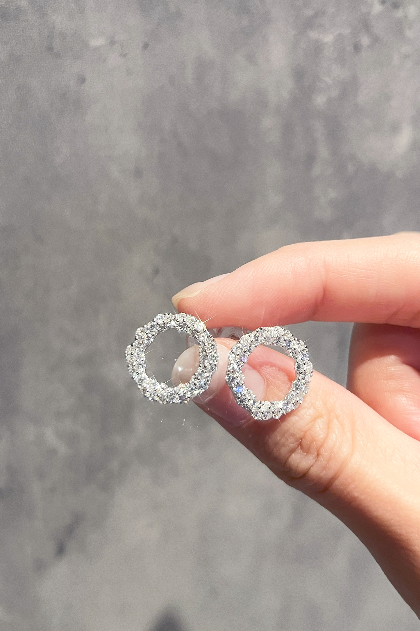 Close-up of a hand holding a pair of sparkling silver circle stud earrings encrusted with crystals, showcased against a textured gray background.